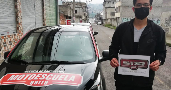 Estudiante graduado con su certificado junto al vehículo de la escuela